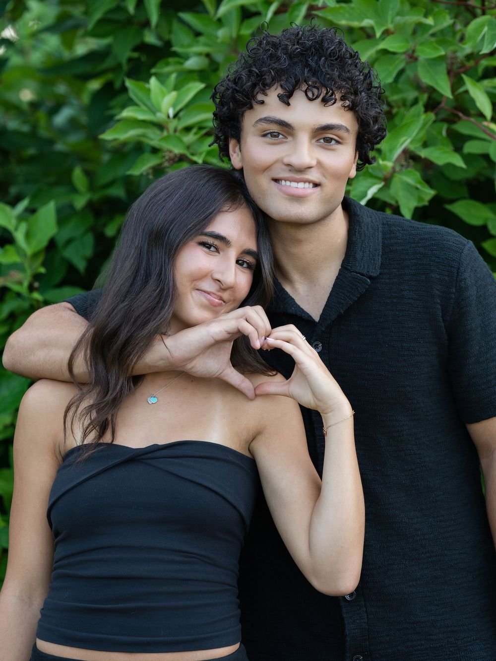 A young couple smiling as the woman makes a heart shape with her hands among green leaves