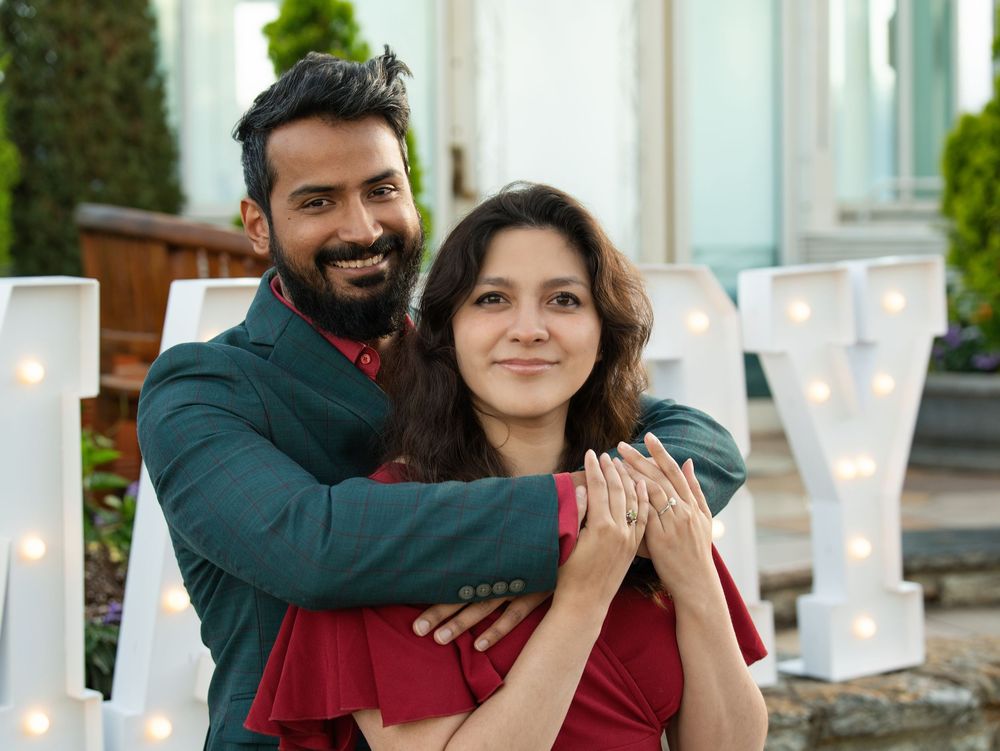 A newly engaged couple embracing in front of lit marquee letters, her ring on display