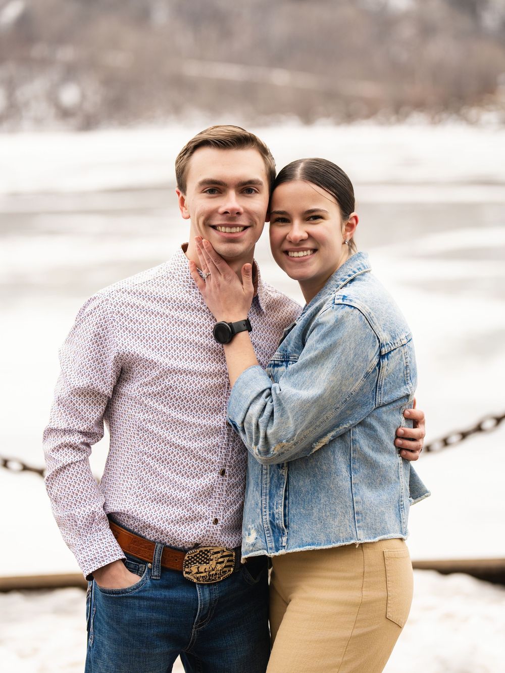 A newly engaged couple smiling cheek to cheek beside an icy river in winter