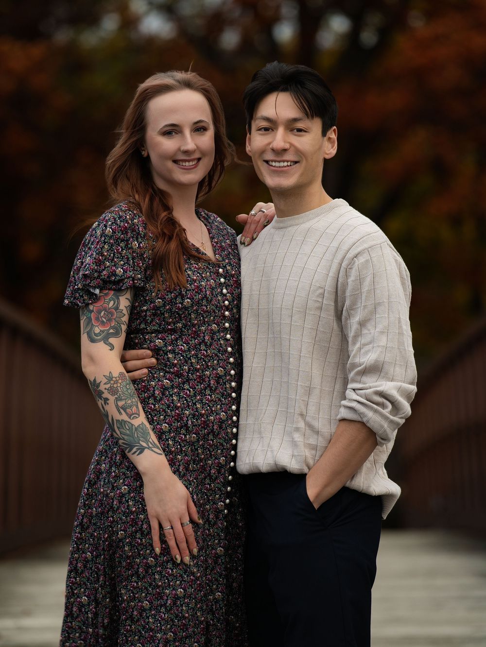 A couple standing arm in arm on a footbridge framed by autumn trees
