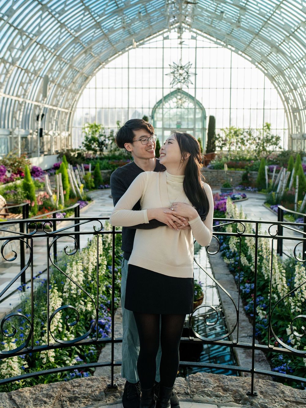 A couple embracing on a walkway inside a glass-domed conservatory full of flowers
