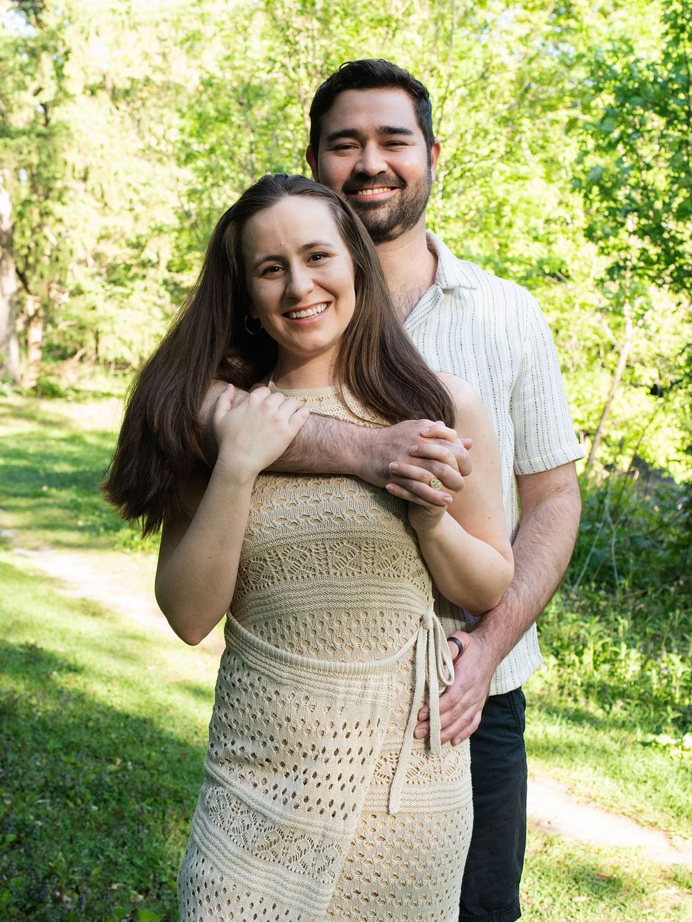 A man hugging a smiling woman from behind on a sunny park trail