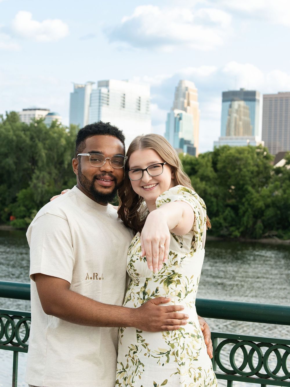 A couple showing off an engagement ring by the river with the Minneapolis skyline behind