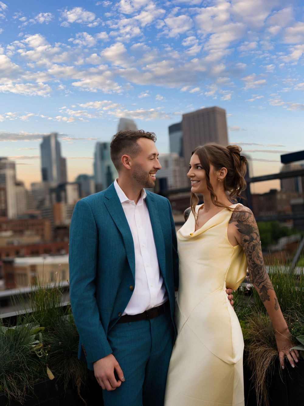 A couple smiling at each other in a rooftop garden with the Minneapolis skyline behind
