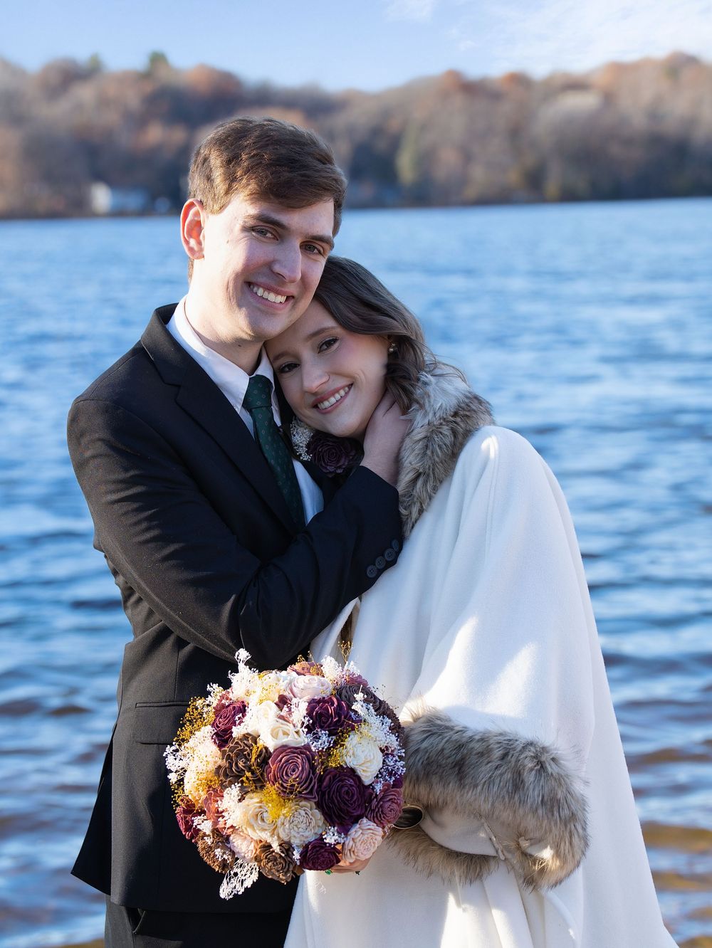 A couple embracing by a lake, the woman in a white cape holding a bouquet