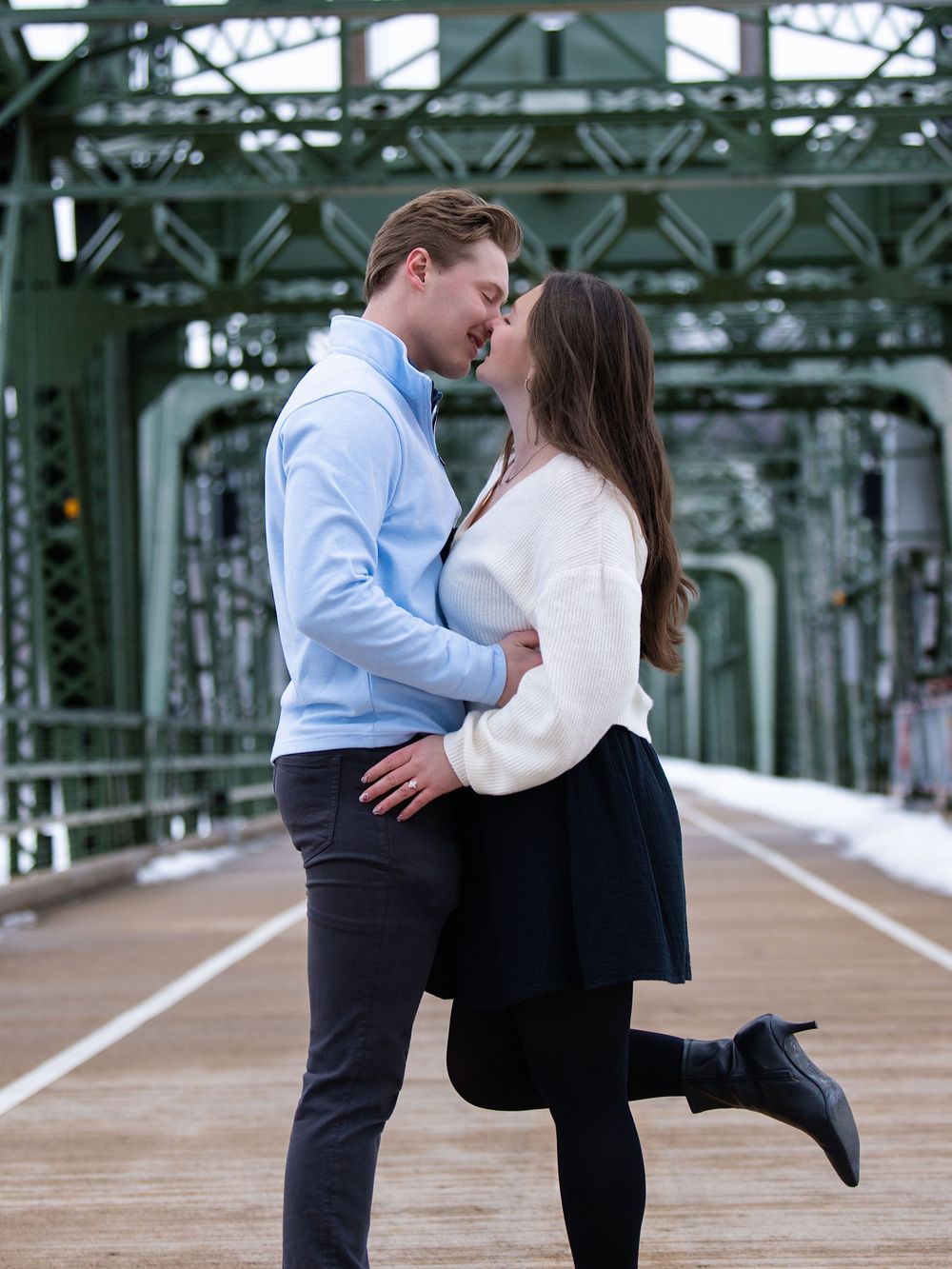 A couple kissing on a green steel truss bridge, the woman lifting one foot