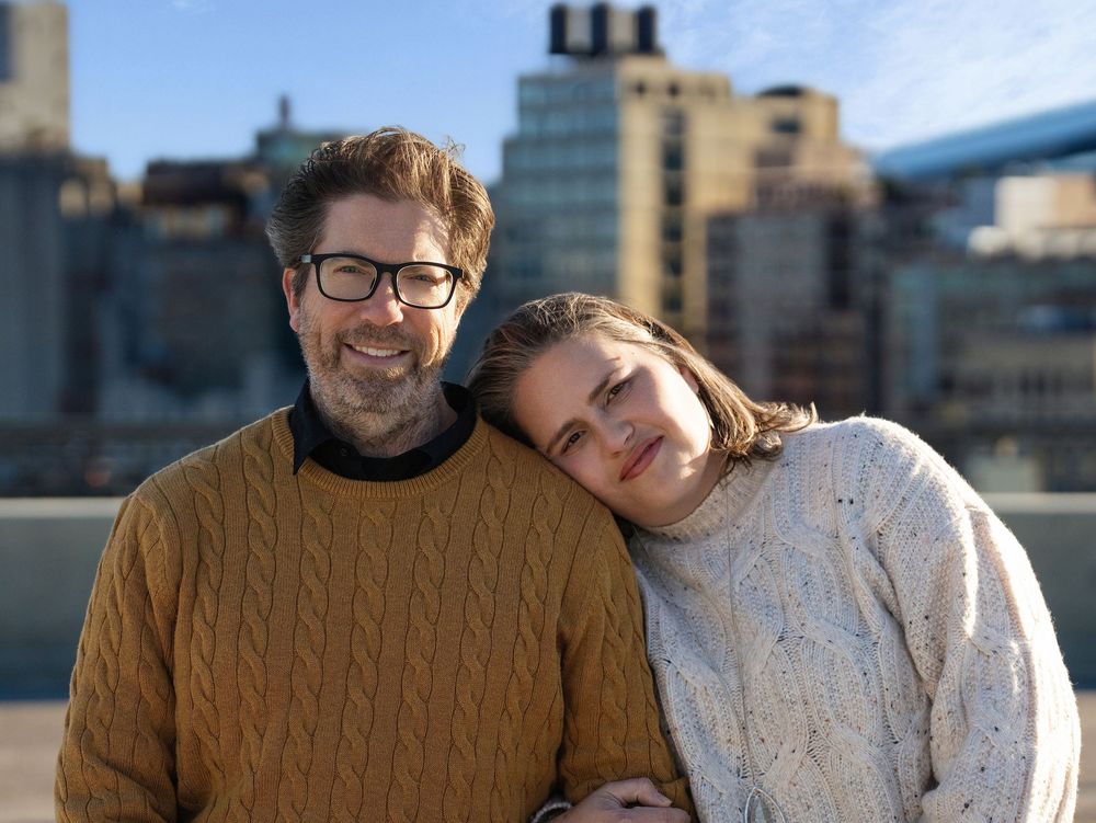 A couple leaning together in cozy sweaters on a rooftop with a city skyline behind