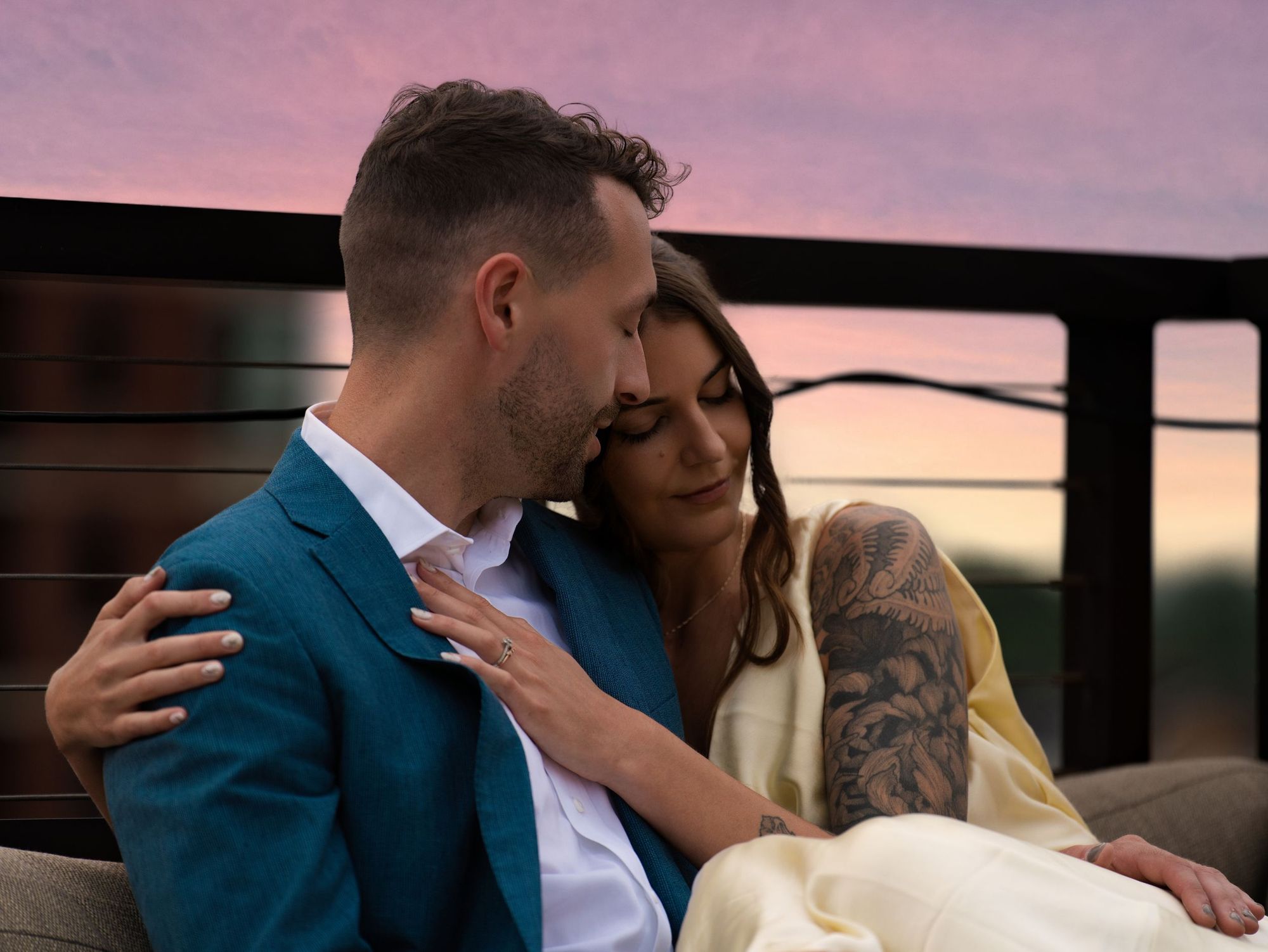 A couple cuddling on a rooftop sofa under a pink sunset sky