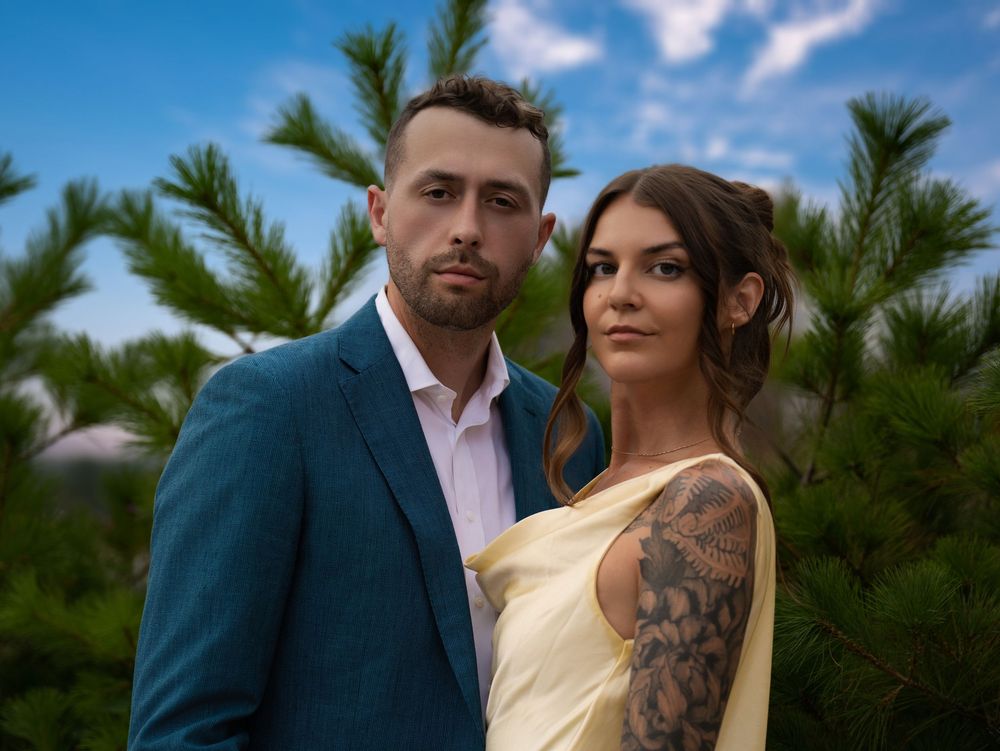 A couple posing among pine trees under a blue sky, the man in a teal suit