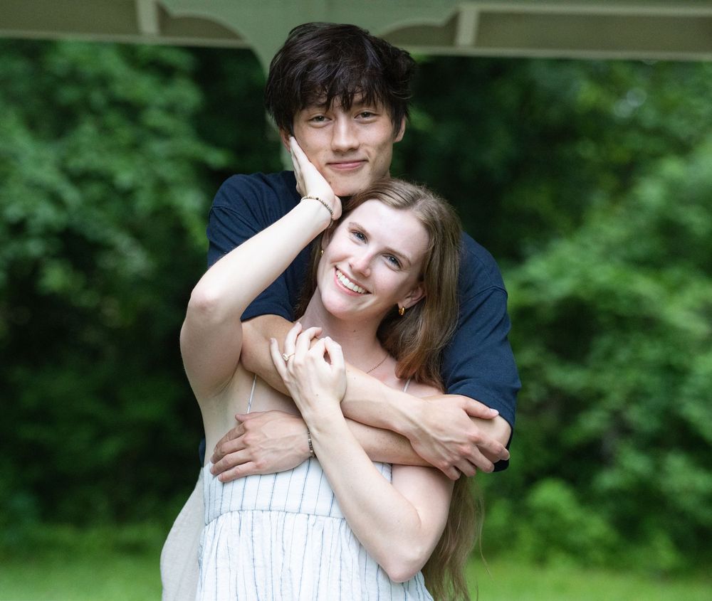 A man hugging a smiling woman from behind as she reaches up to his face in a garden