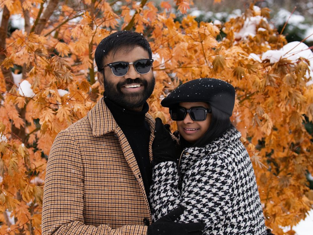 A couple in sunglasses and winter coats smiling among snow-dusted orange autumn leaves