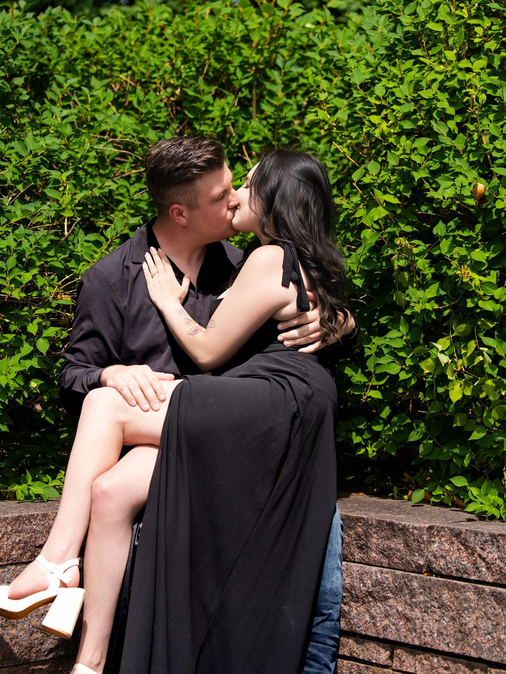 A woman sitting on a man's knee as they kiss in front of a green hedge