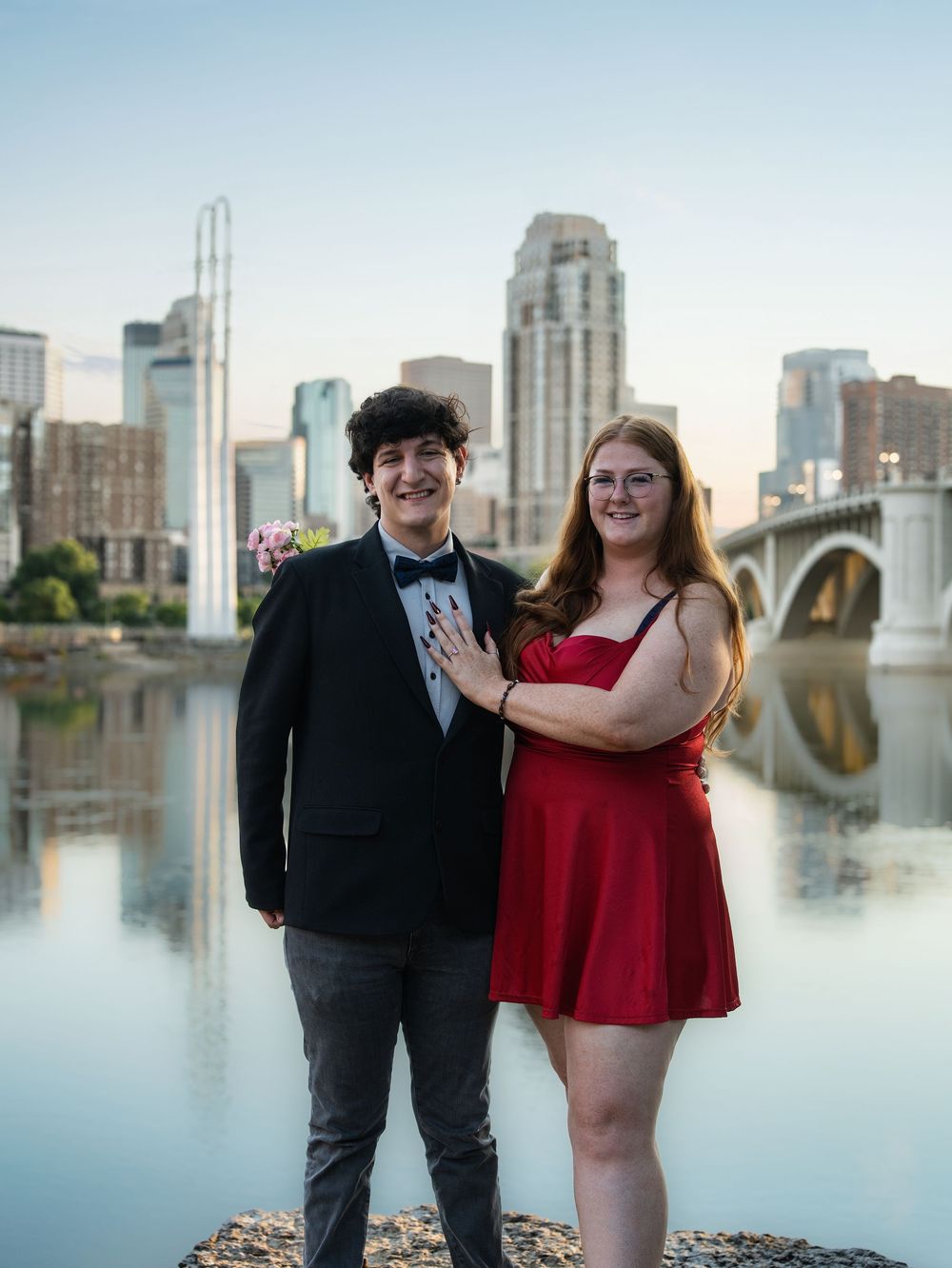 A dressed-up couple standing by the river with the Minneapolis skyline and a bridge behind