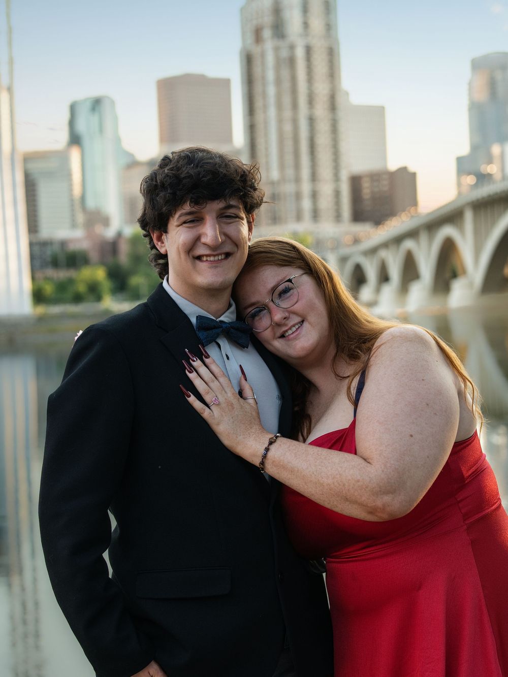 A couple in formal wear embracing by the river with the Minneapolis skyline behind