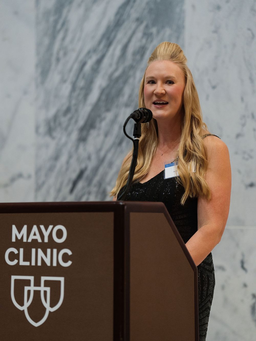A woman speaking at a podium in front of a marble wall at a corporate event