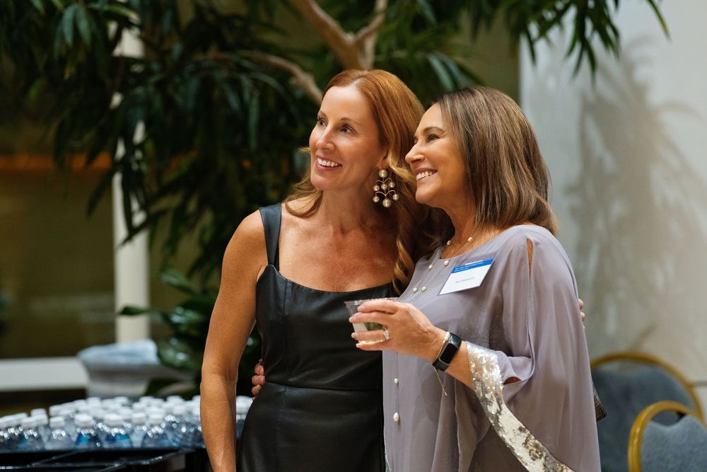 Two women smiling with drinks at a reception in a plant-filled atrium