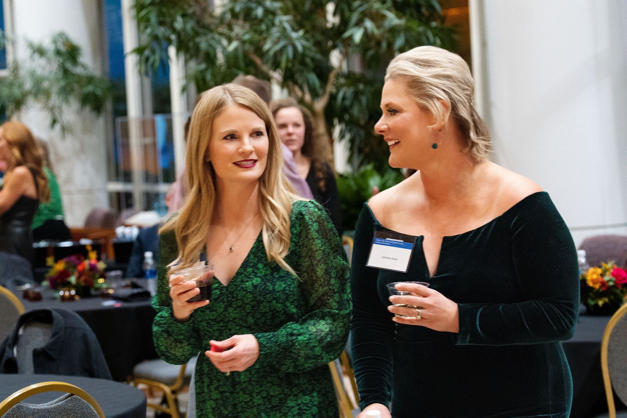 Two women in dark dresses laughing with drinks at a banquet reception