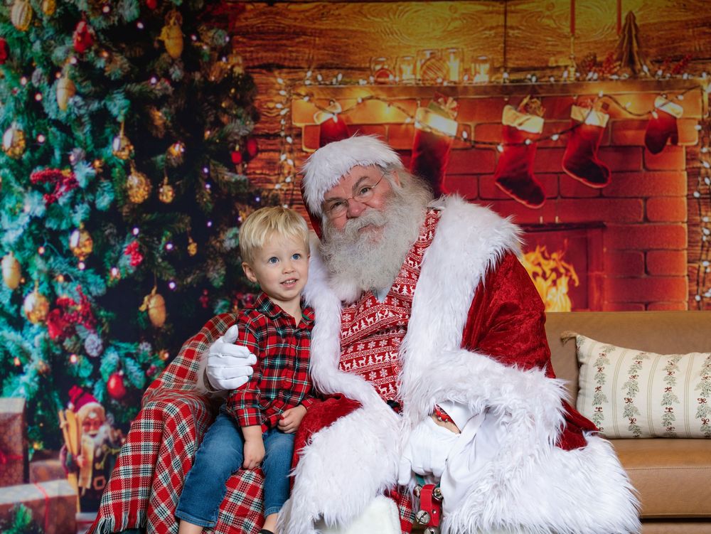 A young boy in a red plaid shirt sitting with Santa Claus beside a festive fireplace