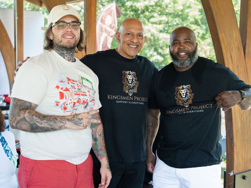 Three men smiling arm in arm under a wooden pavilion at an outdoor community event