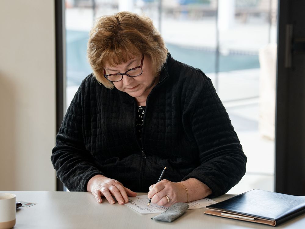 A woman in glasses writing a check at a table beside a large window