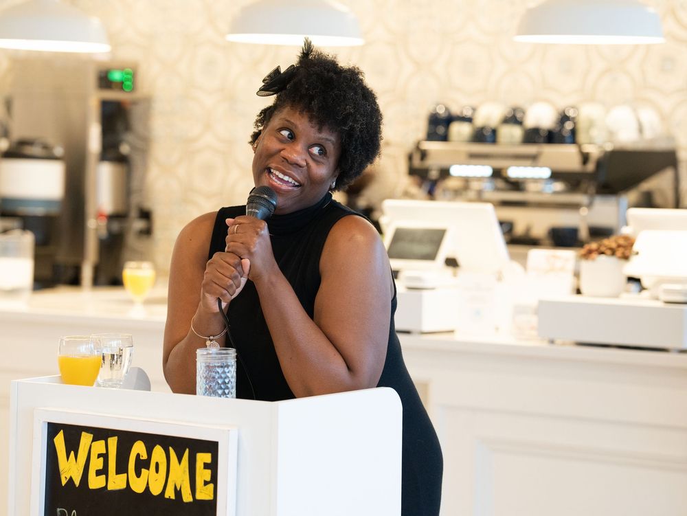 A woman speaking into a microphone behind a welcome sign at an event