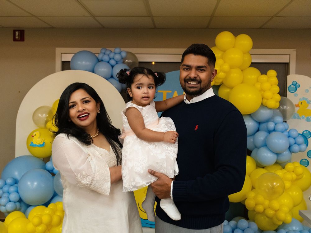 Parents holding their toddler daughter in front of a blue and yellow balloon arch at a party
