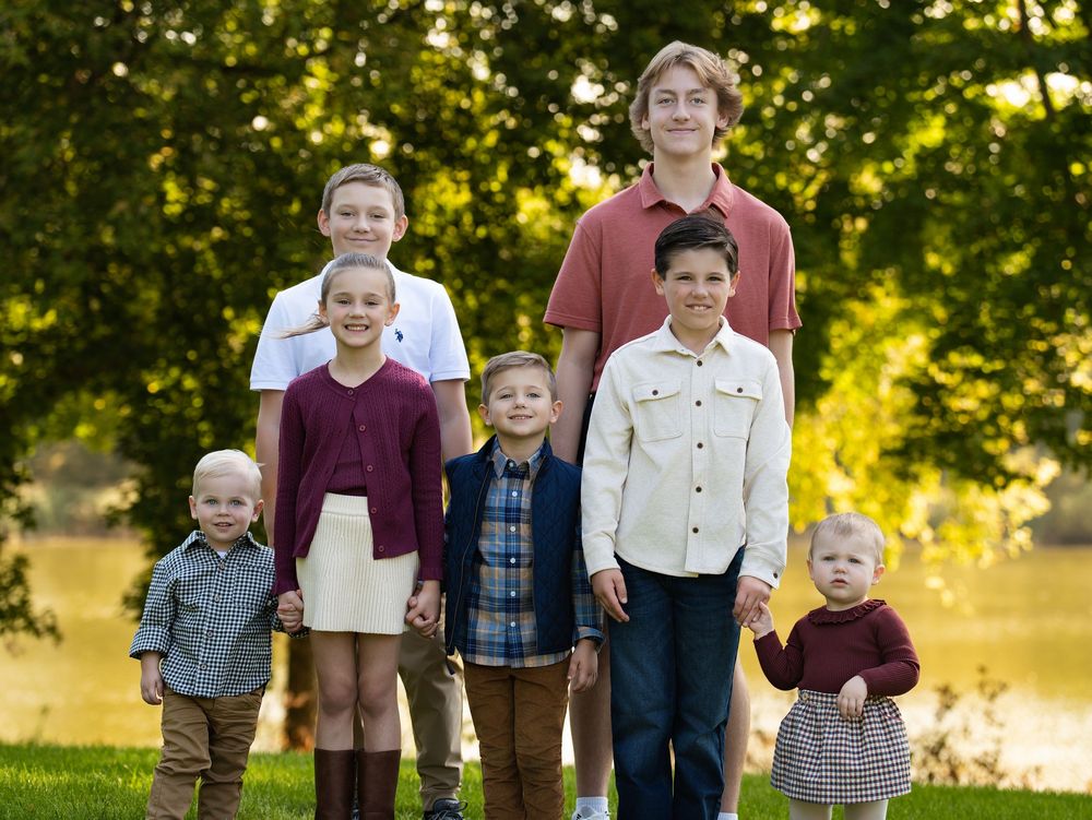 Six children of different ages standing together beside a pond under sunlit trees