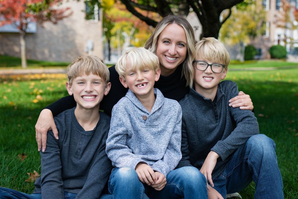 A woman sitting on the grass hugging three smiling boys in a backyard