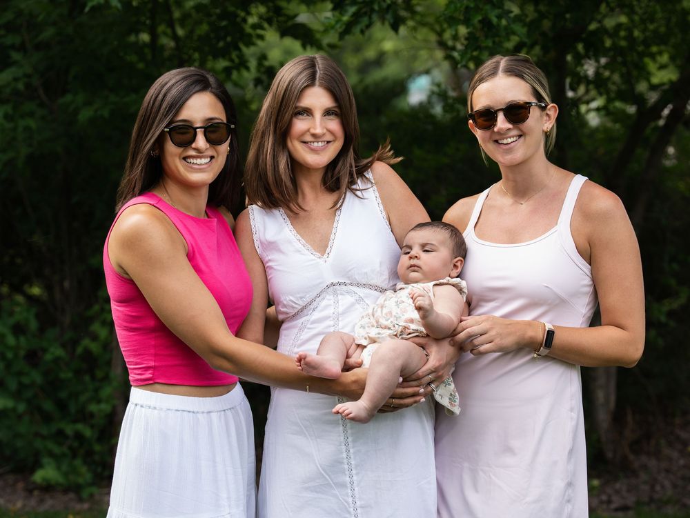 Three women smiling together outdoors, the one in the middle holding a baby