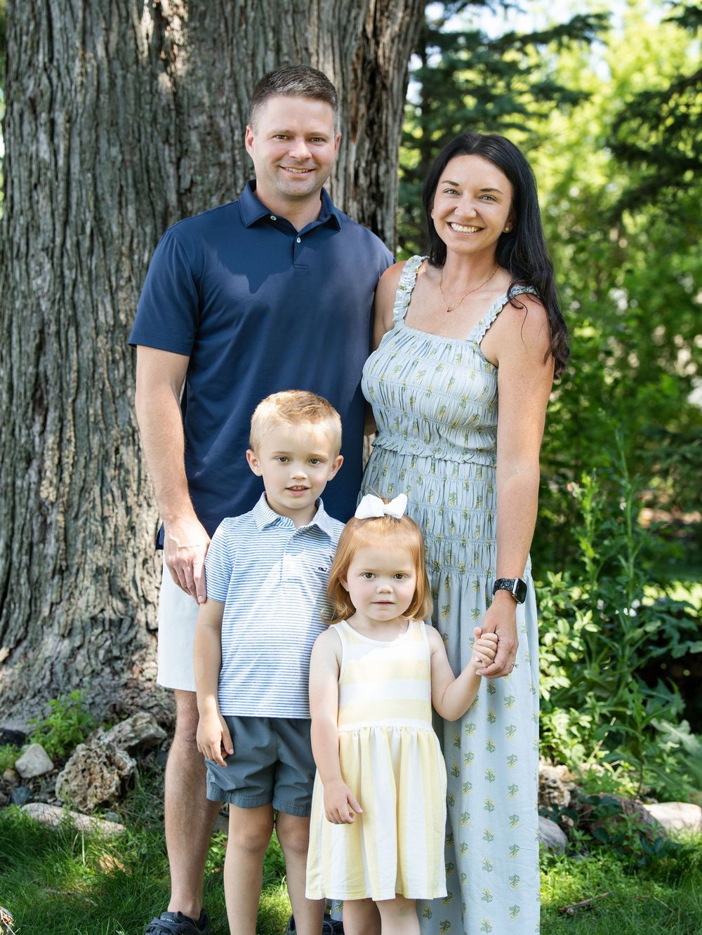 A family of four standing in front of a large tree trunk in summer