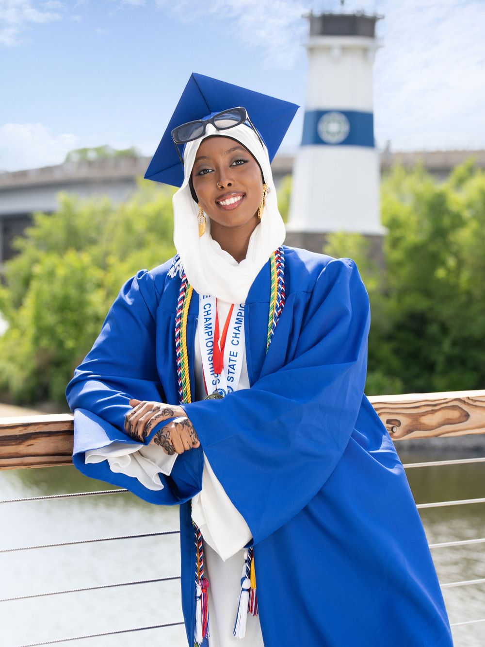 A graduate in a blue gown and white headscarf leaning on a riverside railing near a lighthouse tower