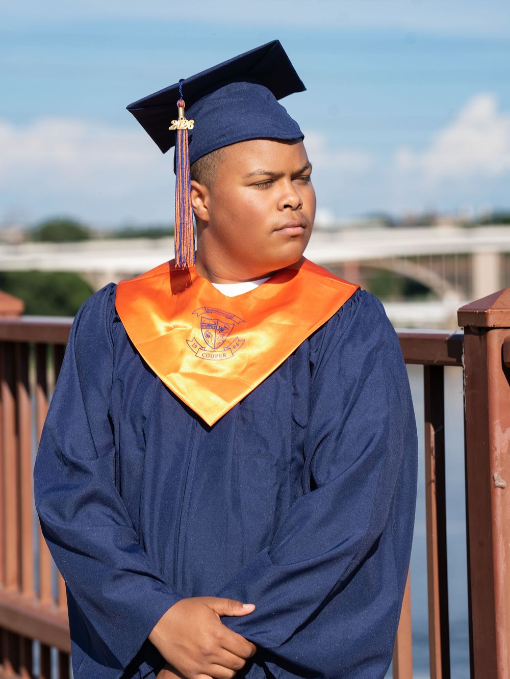 A graduate in a navy gown and orange stole standing on a bridge above a river
