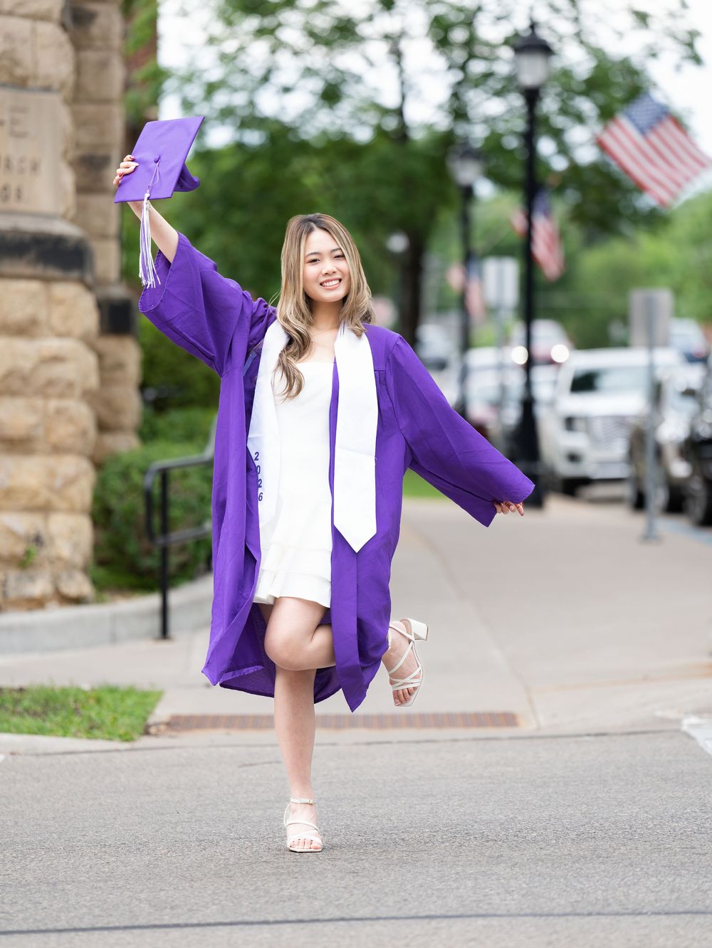 A graduate in a purple gown lifting her cap high while posing on a city sidewalk
