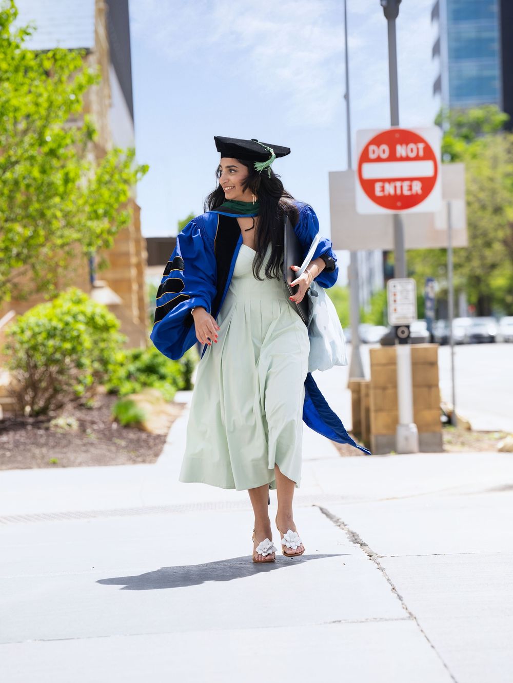 A graduate in a blue doctoral robe and pale green dress walking along a sunny city sidewalk