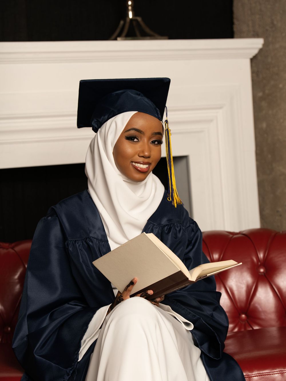 A graduate in a navy gown and headscarf reading a book in a red leather armchair