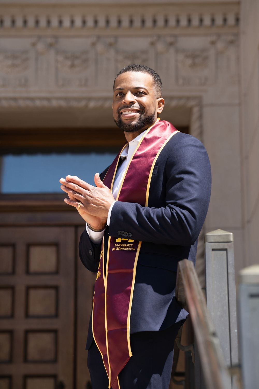 A graduate in a navy suit and maroon stole clapping in front of a carved stone doorway