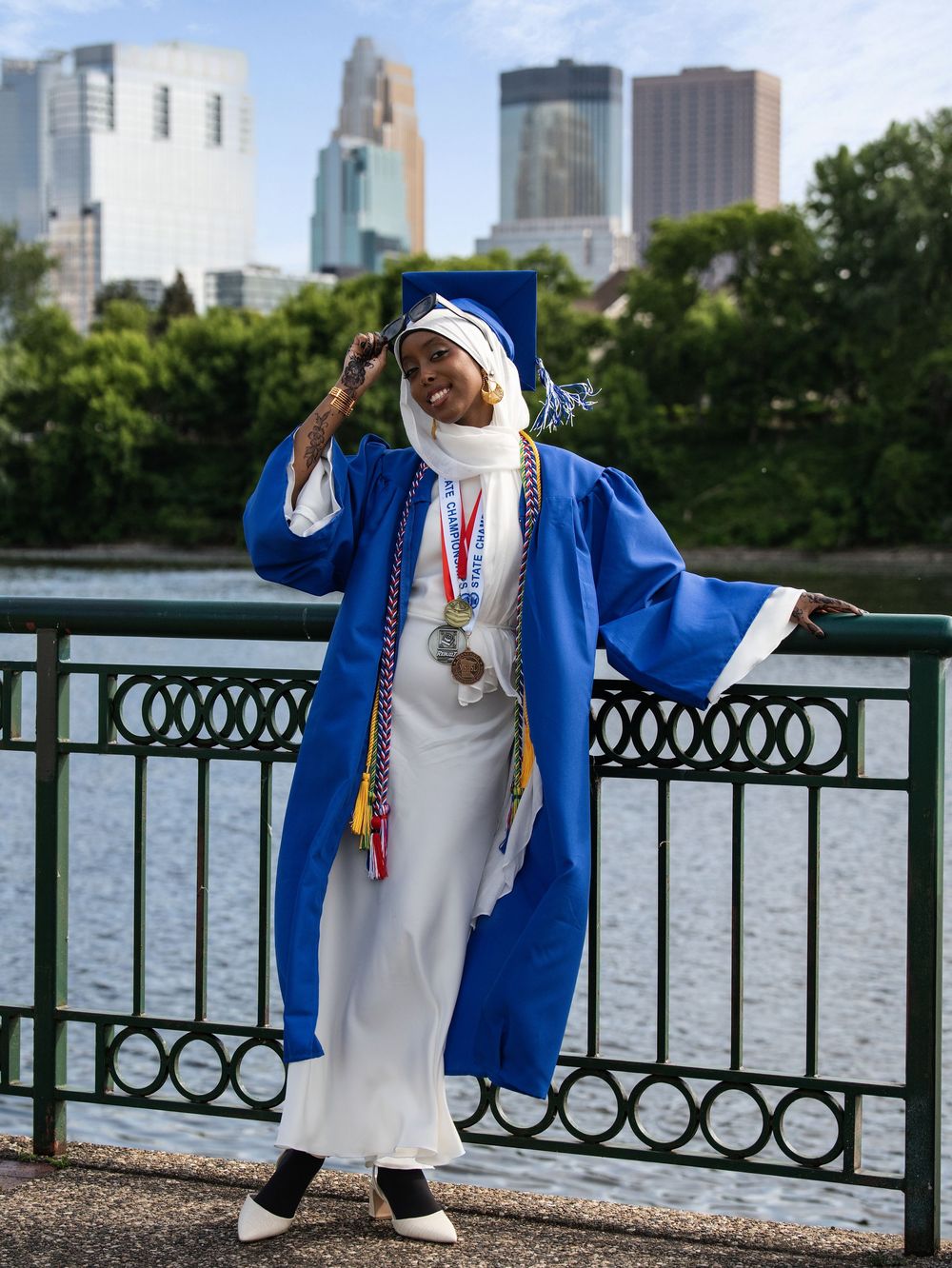 A graduate in a blue gown adjusting her cap at a riverside railing below the Minneapolis skyline