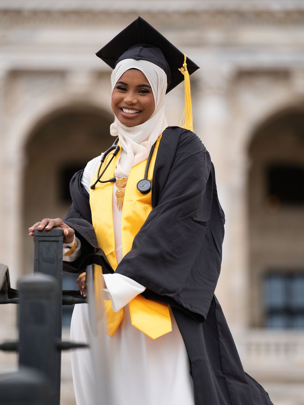 A graduate in a black gown and gold stole with a stethoscope smiling beside stone arches