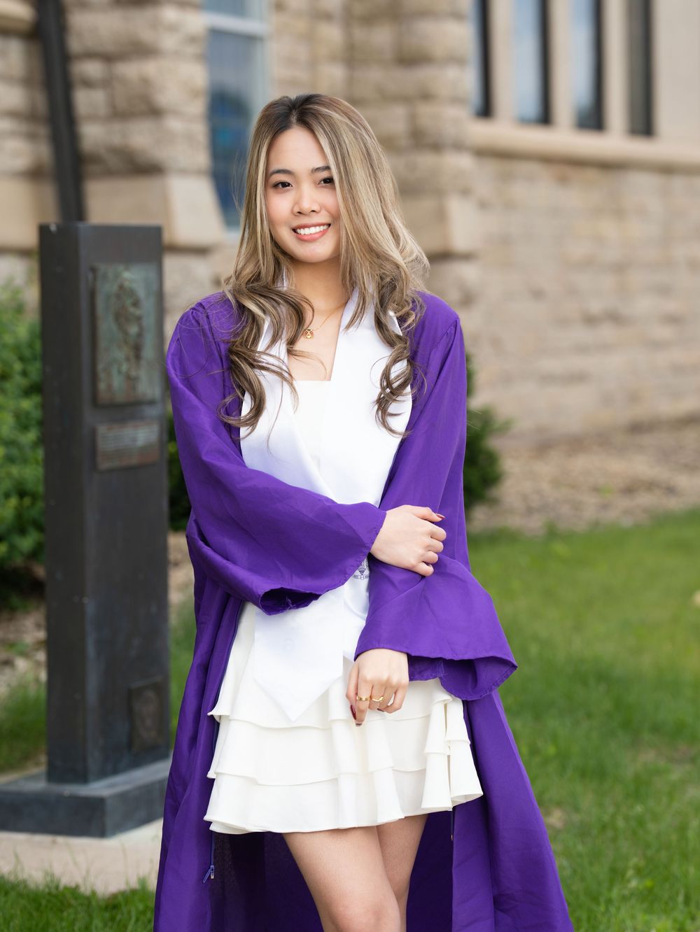 A graduate in a purple gown over a white dress posing beside a stone building