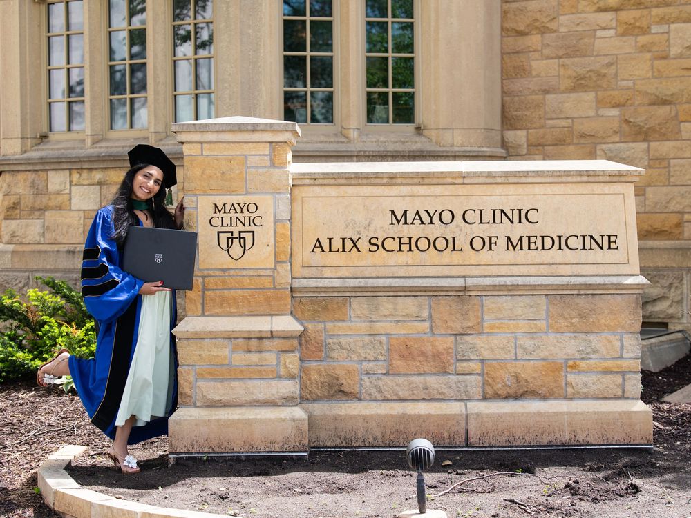 A medical graduate in a doctoral gown posing beside the Mayo Clinic Alix School of Medicine sign