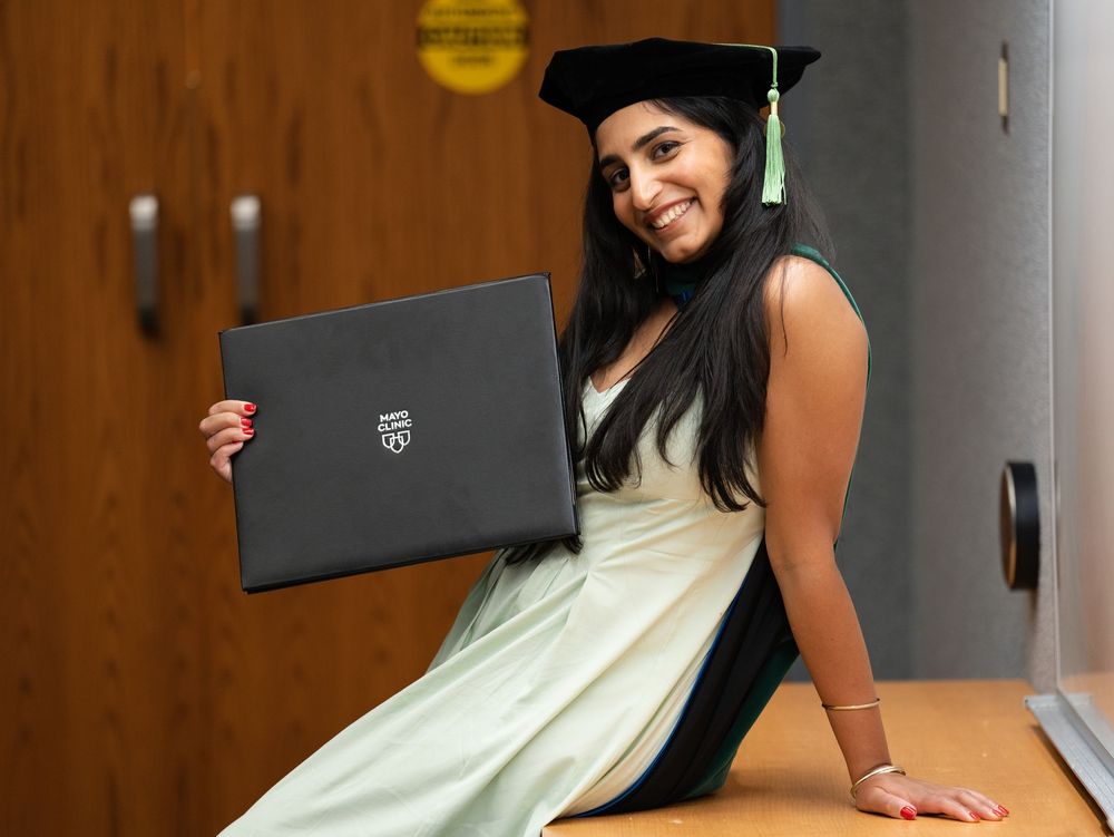A graduate in a doctoral cap sitting on a desk and holding up a diploma cover