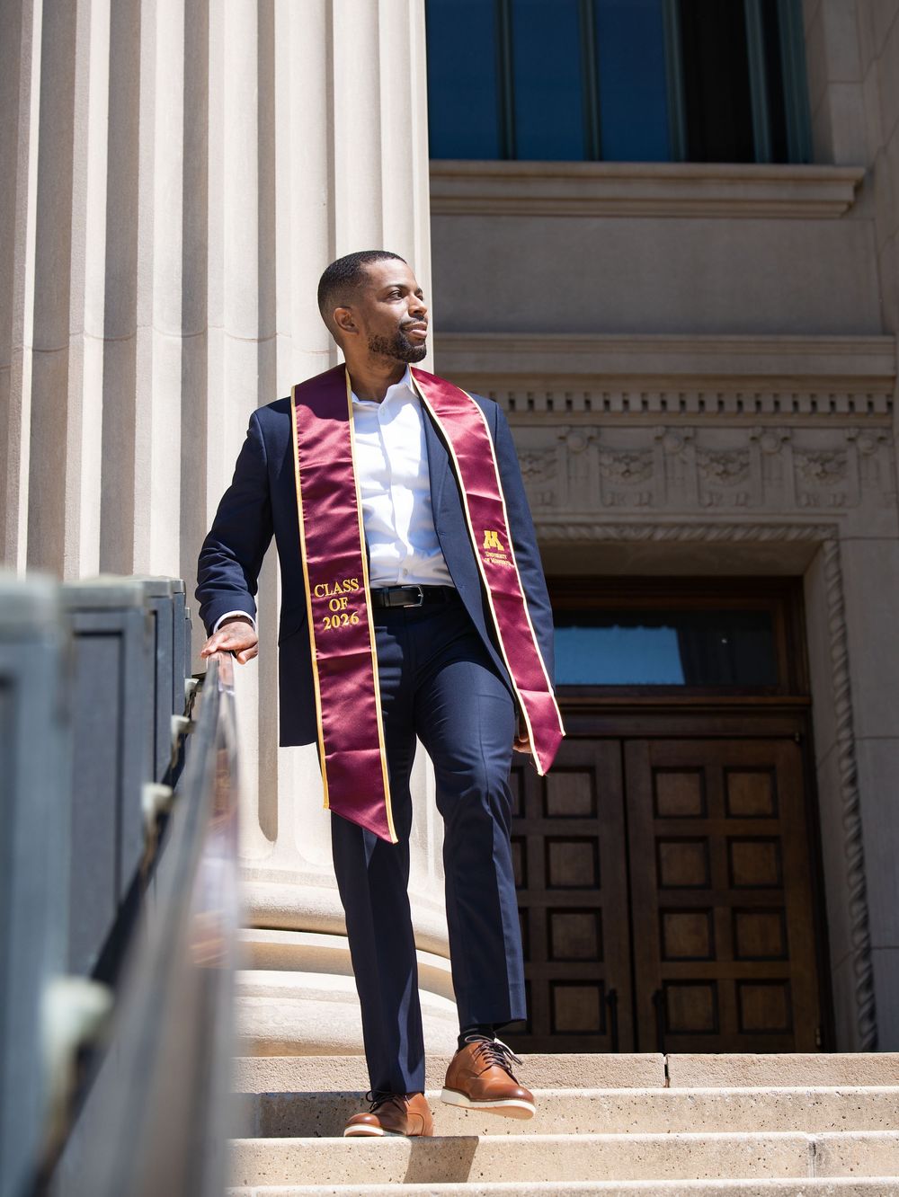 A graduate in a navy suit and maroon stole walking down the steps of a columned building