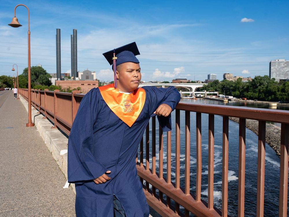 A graduate in a navy gown and orange stole leaning on a bridge railing above a river