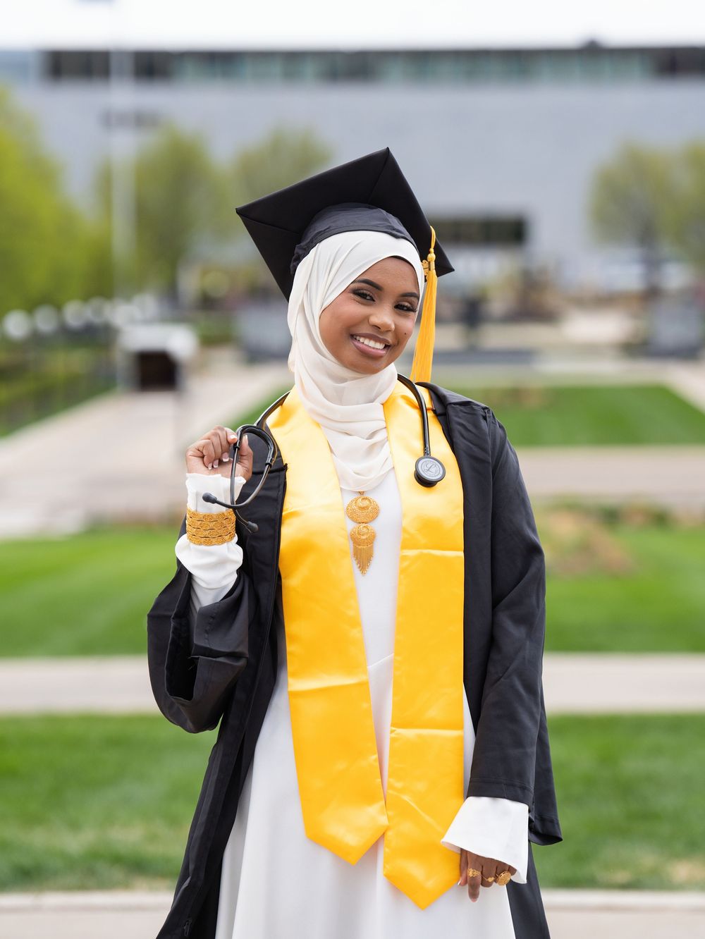 A graduate in a black gown and gold stole holding a stethoscope in a riverside park