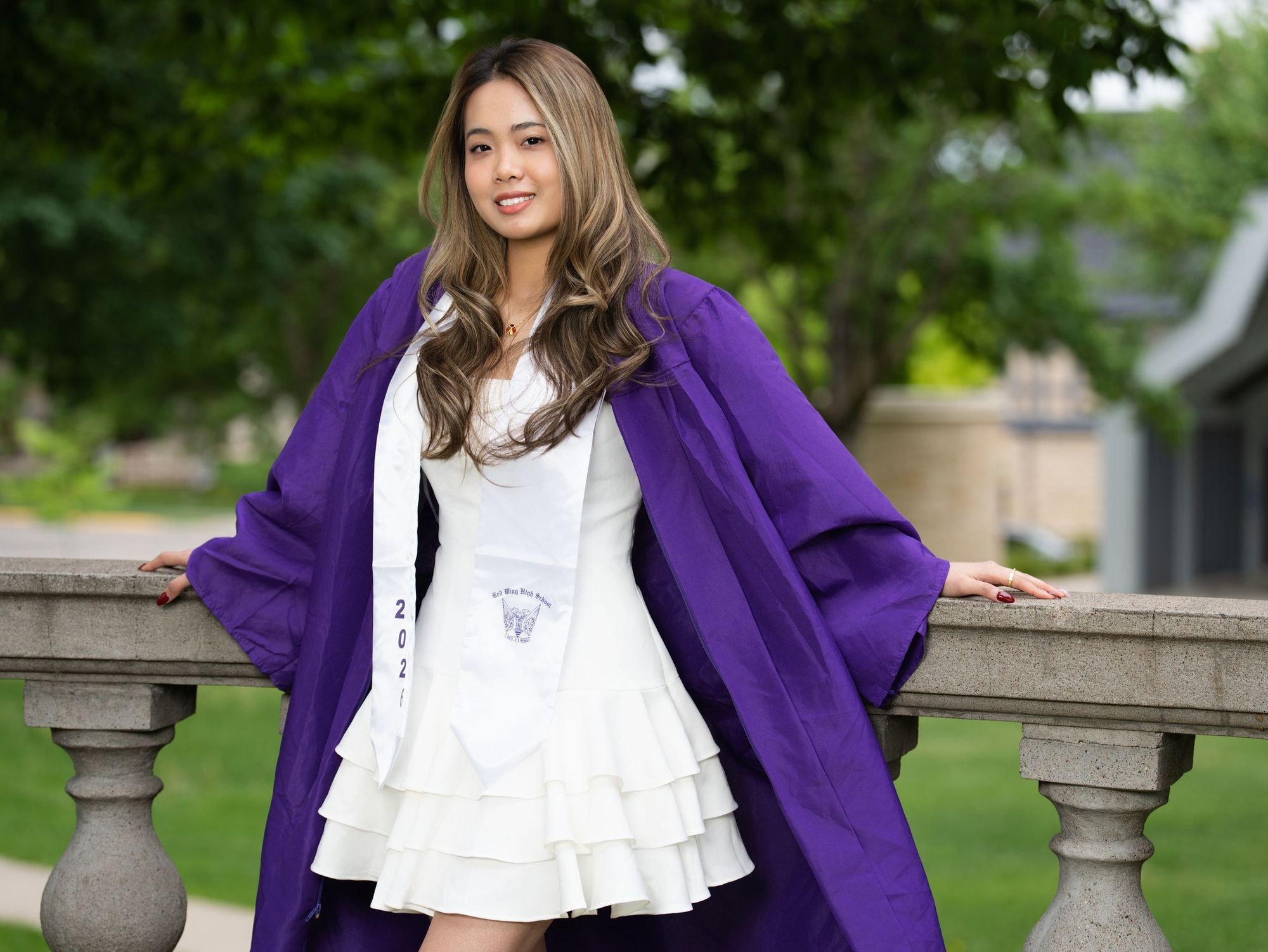 A graduate in a purple gown over a white dress leaning on a stone balustrade in a park
