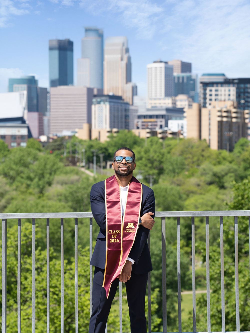 A graduate in a suit and maroon stole at an overlook with the Minneapolis skyline behind