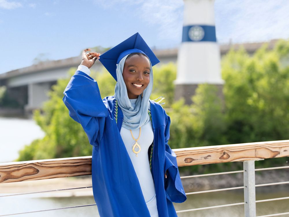 A graduate in a blue gown and headscarf tipping her cap by a river near a lighthouse tower