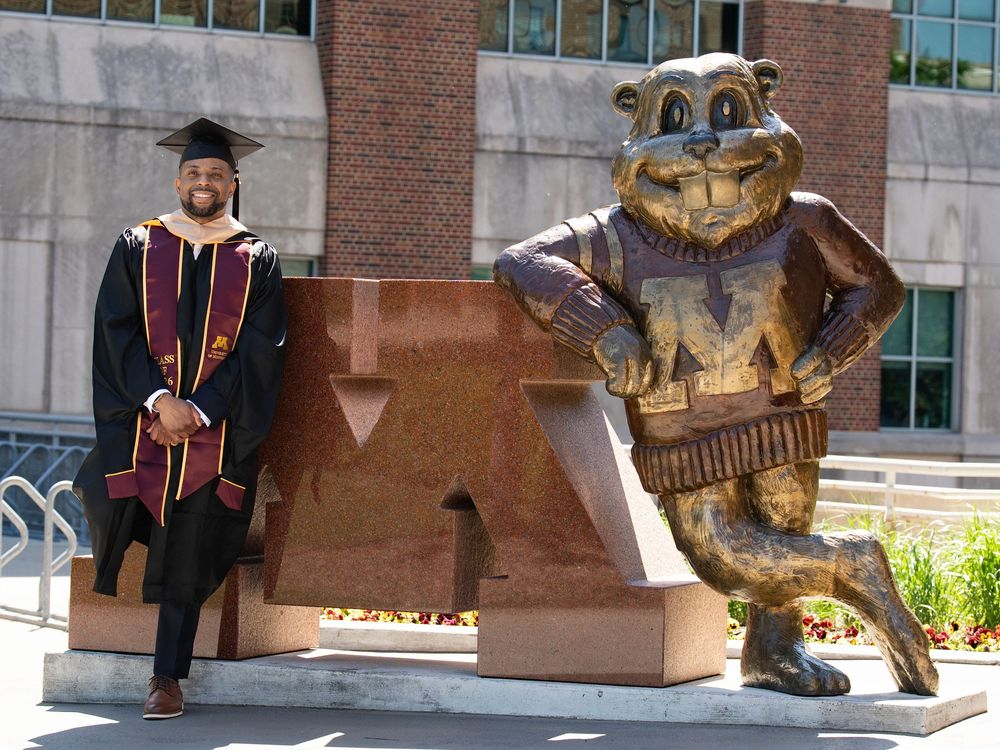 A graduate in a maroon and gold stole beside the University of Minnesota Goldy Gopher statue