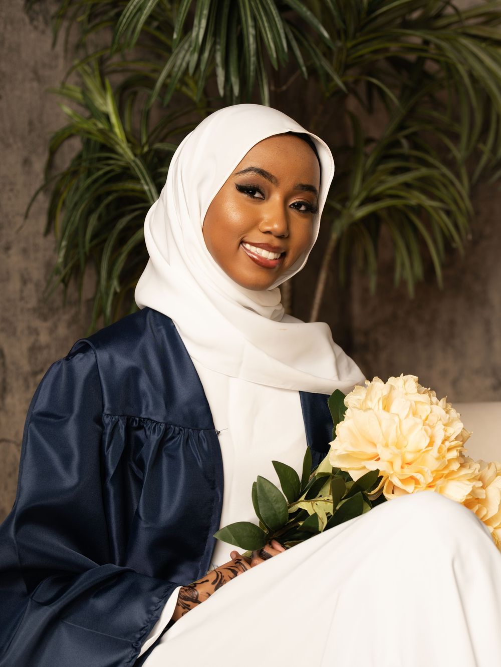A graduate in a navy gown and white headscarf holding cream flowers in front of leafy plants