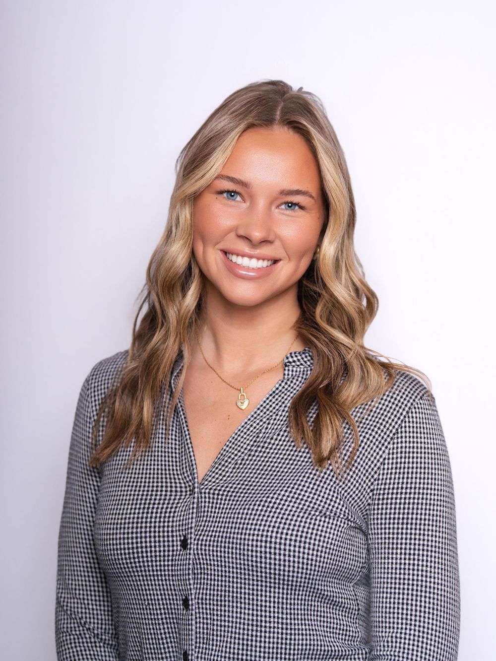 A smiling woman with long wavy hair in a gingham button-up shirt against a light studio backdrop